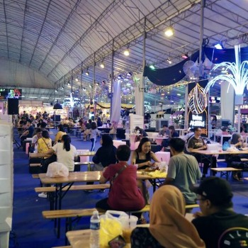 indoor pasar malam night market food court under tent, blue-lit draped ceiling, people seated at long tables eating, festive decorations