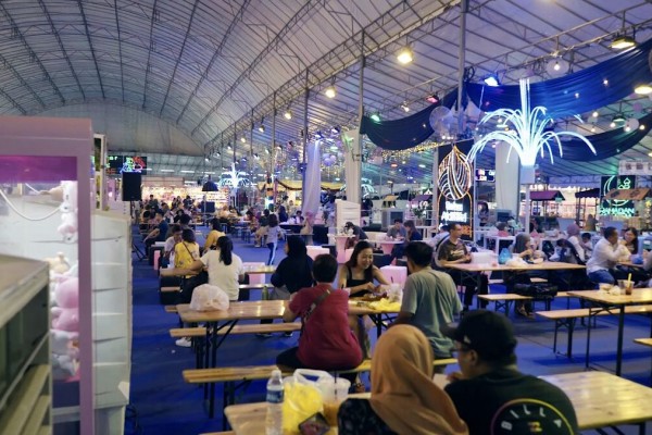 indoor pasar malam night market food court under tent, blue-lit draped ceiling, people seated at long tables eating, festive decorations
