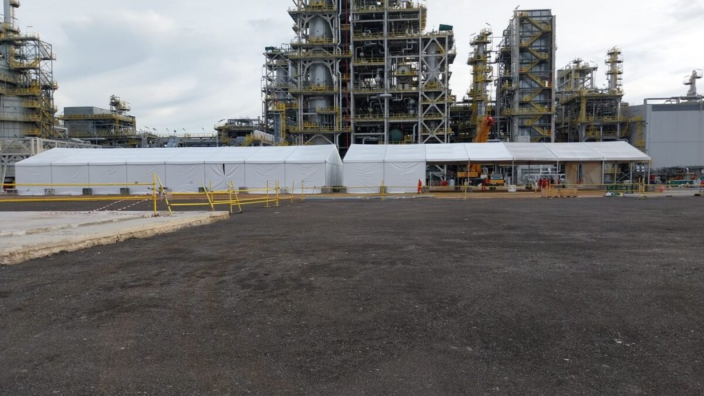 white industrial tent structure with sloped roof in front of large petrochemical refinery towers, wide panoramic shot
