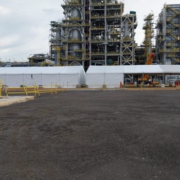 white industrial tent structure with sloped roof in front of large petrochemical refinery towers, wide panoramic shot