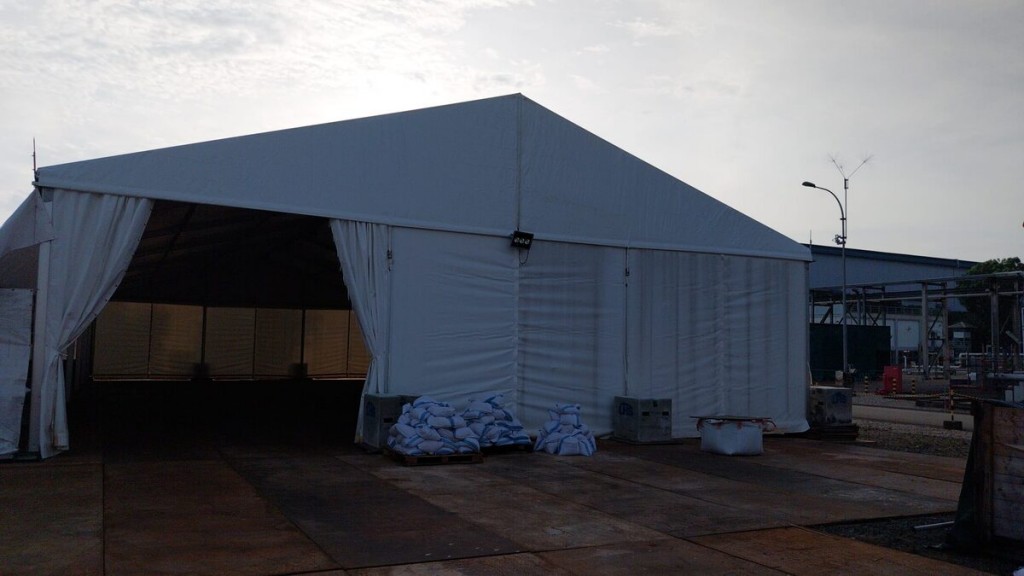 white A-frame tent exterior at industrial site, sandbags weighing down base, overcast sky, industrial buildings and streetlight in background