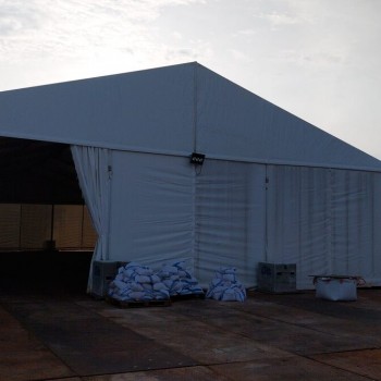white A-frame tent exterior at industrial site, sandbags weighing down base, overcast sky, industrial buildings and streetlight in background