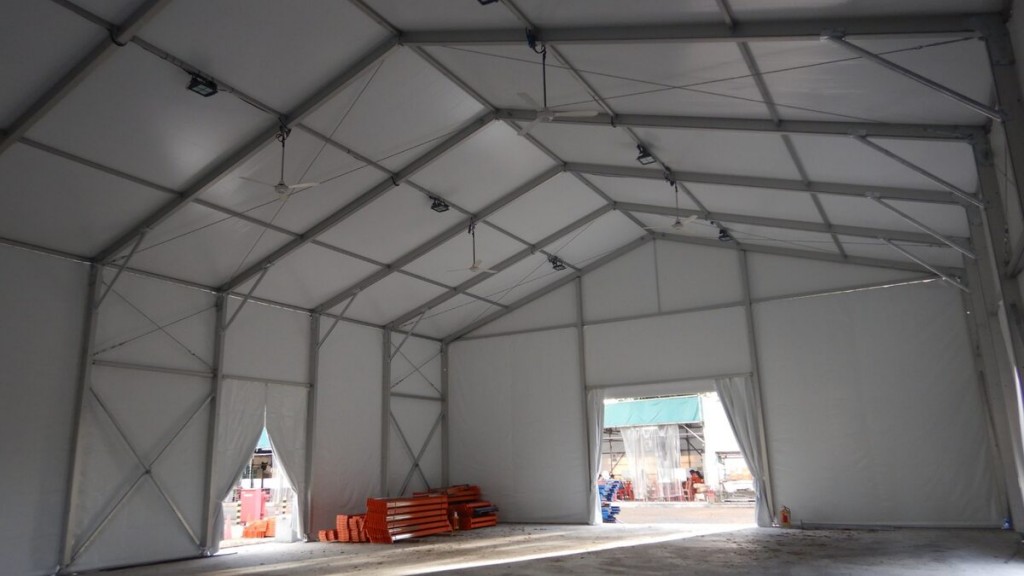 interior of empty white tent, exposed truss ceiling, storage shelving racks visible through opening, concrete floor, industrial setting