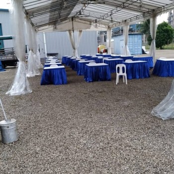 white tent with exposed frame ceiling, blue-skirted rectangular tables, outdoor gravel area, industrial building backdrop, empty