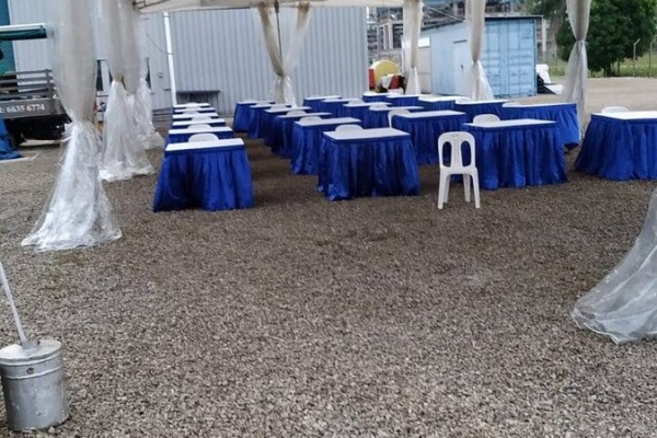 white tent with exposed frame ceiling, blue-skirted rectangular tables, outdoor gravel area, industrial building backdrop, empty