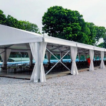 outdoor daytime white marquee tent with draped side curtains, rows of black chairs, gravel ground, trees in background