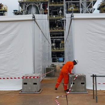white tent gap between structures at industrial plant, worker in orange coveralls kneeling at concrete blocks with safety tape, pipework visible