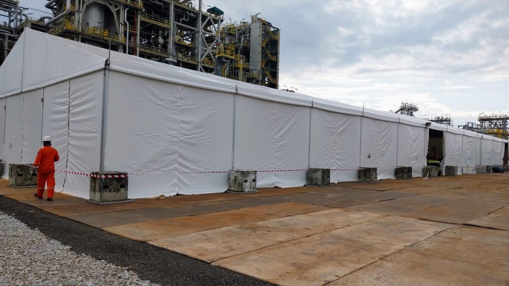 large white industrial tent at petrochemical plant site, worker in orange coveralls and hard hat standing outside, safety barrier tape, industrial pipes in background