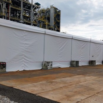 large white industrial tent at petrochemical plant site, worker in orange coveralls and hard hat standing outside, safety barrier tape, industrial pipes in background