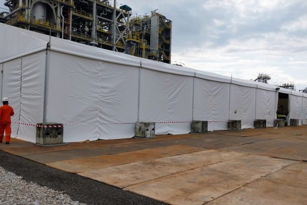 large white industrial tent at petrochemical plant site, worker in orange coveralls and hard hat standing outside, safety barrier tape, industrial pipes in background