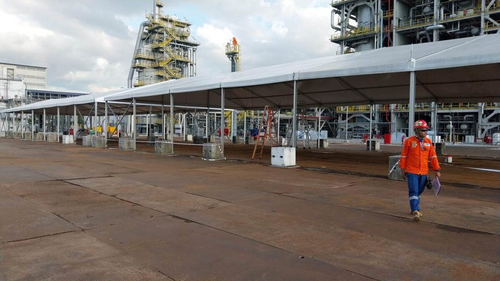 white sloped tent roof structure next to large petrochemical plant facility, worker in orange coveralls, industrial pipes and towers