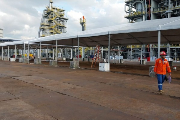 white sloped tent roof structure next to large petrochemical plant facility, worker in orange coveralls, industrial pipes and towers