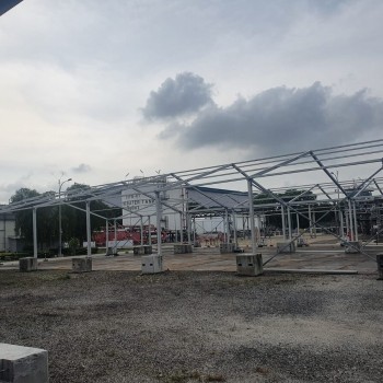 aluminium tent frame skeleton being erected at industrial plant site, concrete ballast blocks, no walls yet, overcast sky