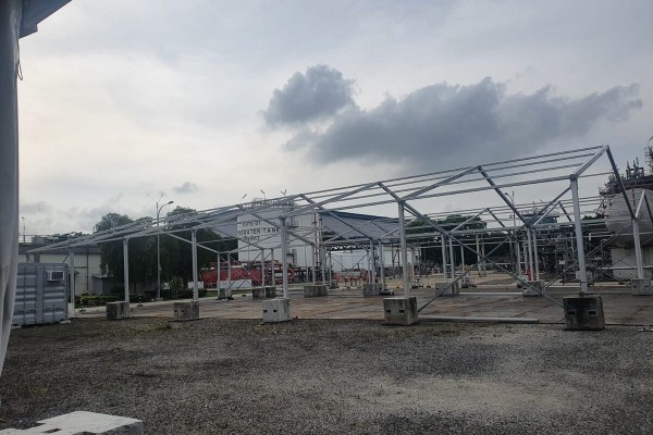aluminium tent frame skeleton being erected at industrial plant site, concrete ballast blocks, no walls yet, overcast sky