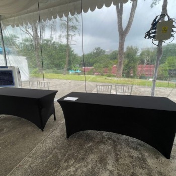 outdoor black-clothed rectangular tables under tent edge, portable aircon unit beside table, cloudy sky, grass and trees in background