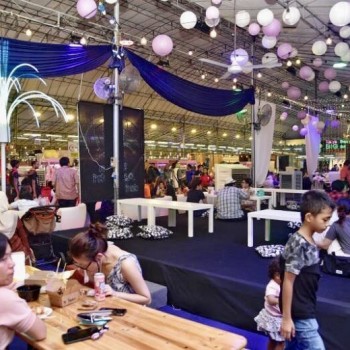 indoor pasar malam bazaar with purple and white balloon decorations, string lights, families seated at long tables, festive atmosphere